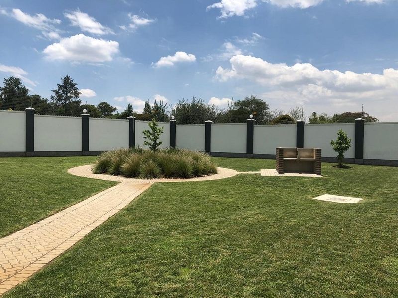 A well-maintained garden featuring a paved walkway, a central landscaped bed, and a built-in brick braai, enclosed by a secure perimeter wall.