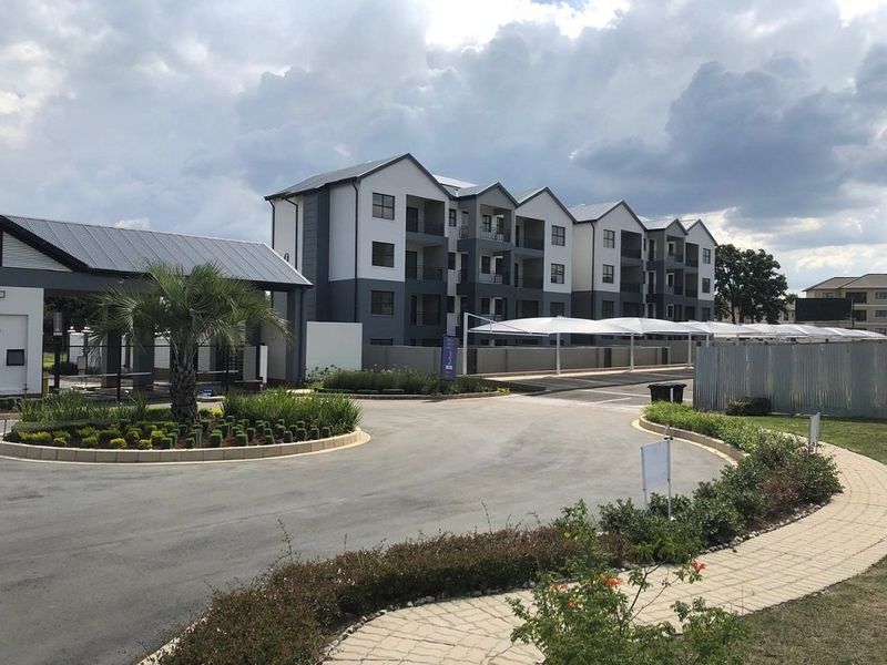 A view of the apartment complex entrance featuring a paved driveway, landscaped gardens, and covered parking bays under shade cloth structures.