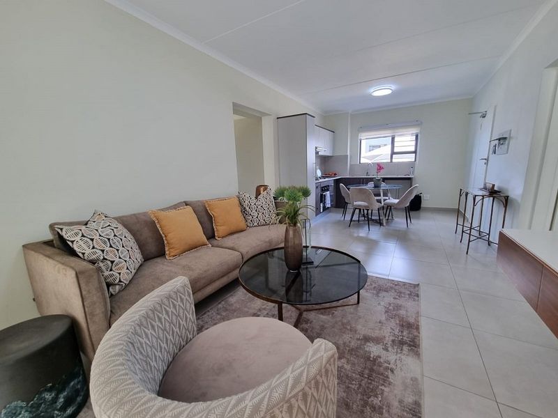 An open-plan living and kitchen area featuring neutral tiled flooring, modern cabinetry, and integrated kitchen appliances in a well-maintained apartment setting.
