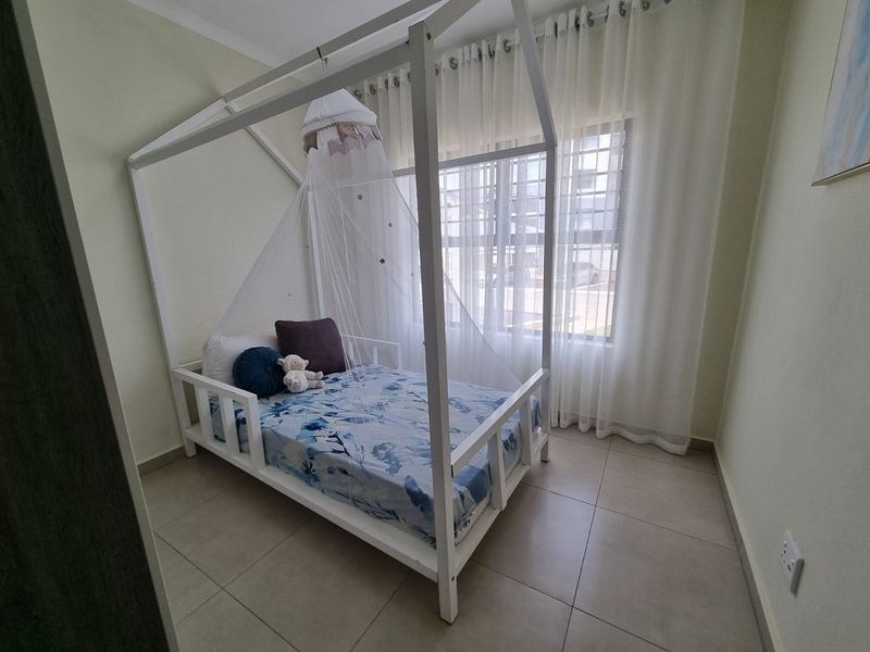 A bright bedroom featuring neutral tiled flooring and a large window fitted with security bars and sheer curtains.