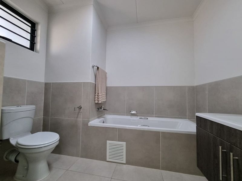 A clean, modern bathroom featuring a built-in bathtub, wall-mounted toilet, and a vanity cabinet with neutral ceramic tiling throughout.