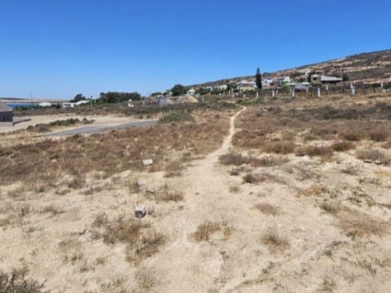 A view of a vacant residential plot featuring natural vegetation and an unpaved access path, situated in a developing coastal neighborhood.