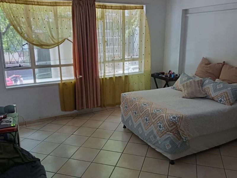 A spacious bedroom featuring tiled flooring and a large window with security bars, offering natural light and a functional layout.