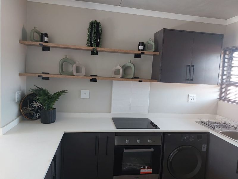 Modern kitchen featuring dark cabinetry, white stone countertops, built-in oven, electric hob, and integrated washing machine with stylish floating shelving.