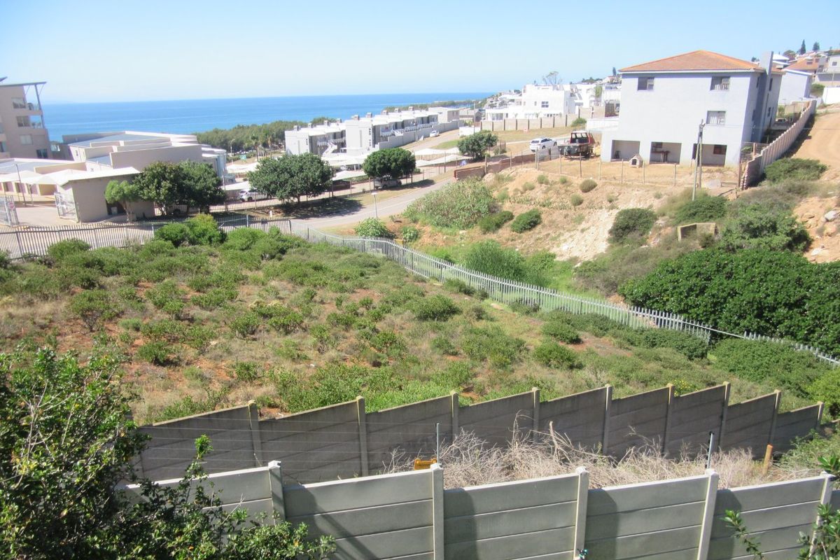 Elevated view of a vacant residential plot featuring established vegetation, enclosed by precast concrete walls and metal palisade fencing, overlooking the coastal neighborhood.