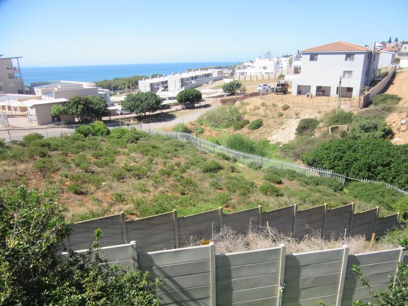Elevated view of a vacant residential plot featuring established vegetation, enclosed by precast concrete walls and metal palisade fencing, overlooking the coastal neighborhood.