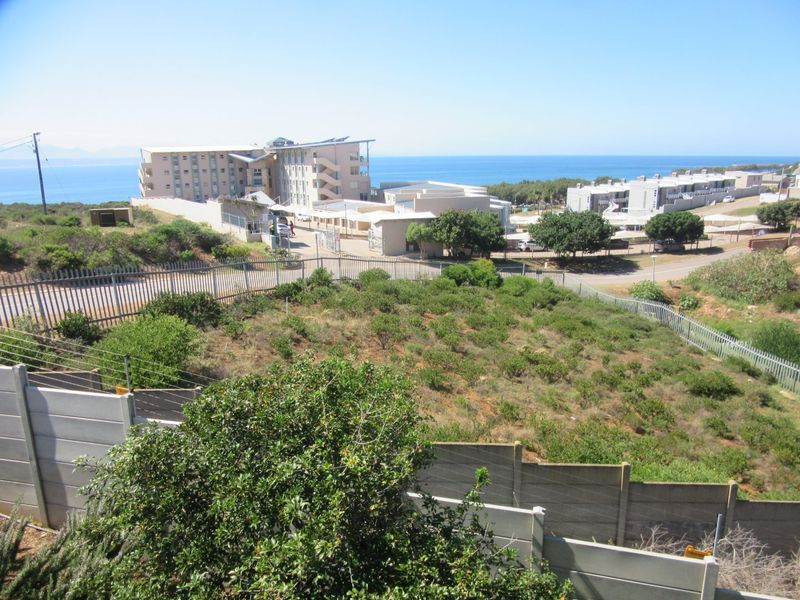 An elevated view of a vacant residential plot featuring perimeter fencing and a retaining wall, with coastal buildings and ocean views in the background.