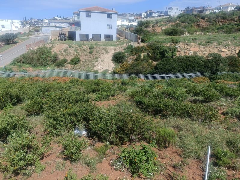 A view of a vacant residential plot featuring sloped terrain and perimeter fencing, situated in a developed suburban neighborhood.