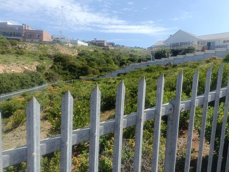 A view of a sloped residential vacant plot featuring a galvanized steel palisade fence and a tiered retaining wall structure.
