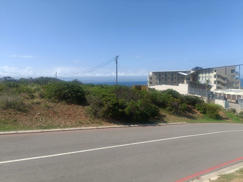 A street-level view of a vacant residential plot featuring coastal vegetation, utility infrastructure, and a view towards the ocean.