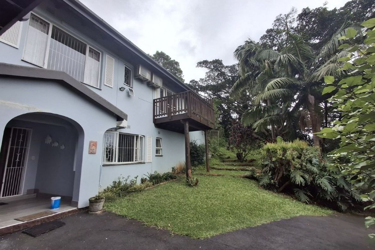 Rear view of the property featuring a raised timber balcony, covered patio area, and established garden with paved driveway access.