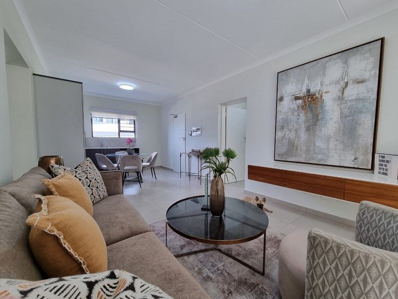 A bright open-plan living and dining area featuring tiled floors, a floating media console, and a functional kitchen space in the background.