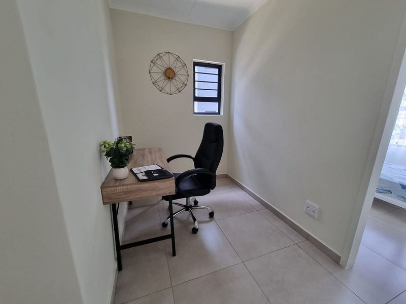 A compact home office space featuring neutral tiled flooring, clean white walls, and a window with security bars.