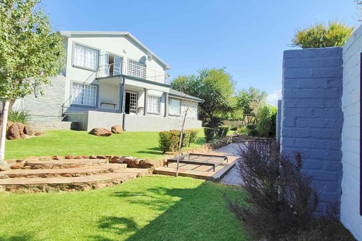 Rear view of the property featuring a landscaped garden, stone retaining walls, a paved patio area, and an upper-level balcony.