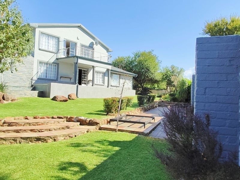 Rear view of the property featuring a landscaped garden, stone retaining walls, a paved patio area, and an upper-level balcony.