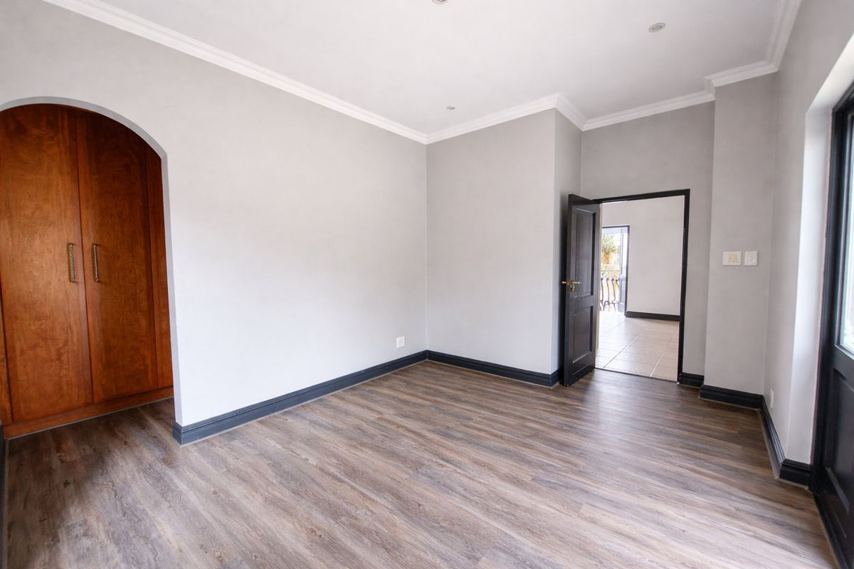A bright, empty bedroom featuring modern laminate flooring, a built-in wooden wardrobe, an elegant archway, and neutral grey walls with dark trim.