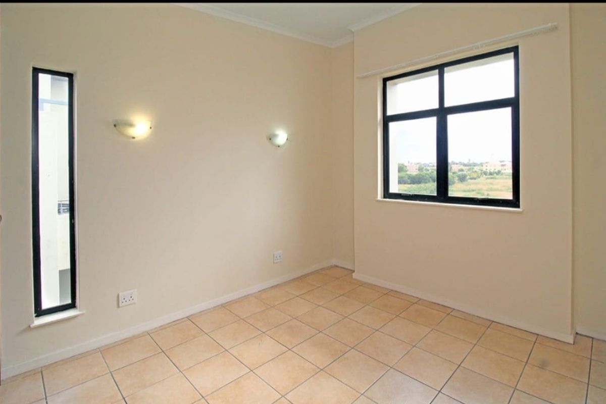 A bright, empty bedroom featuring neutral tiled flooring, black-framed windows, and wall-mounted lighting, offering a clean canvas for personalization.