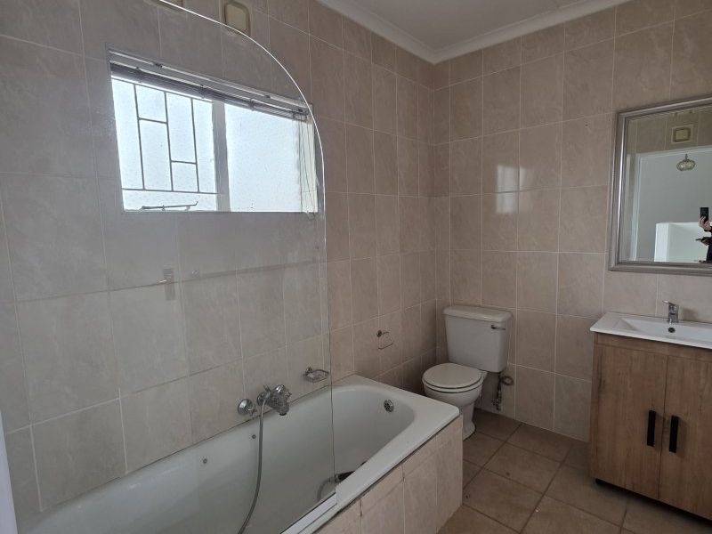 A functional bathroom featuring a built-in bathtub with a glass screen, a vanity cabinet with a basin, and neutral wall and floor tiling.