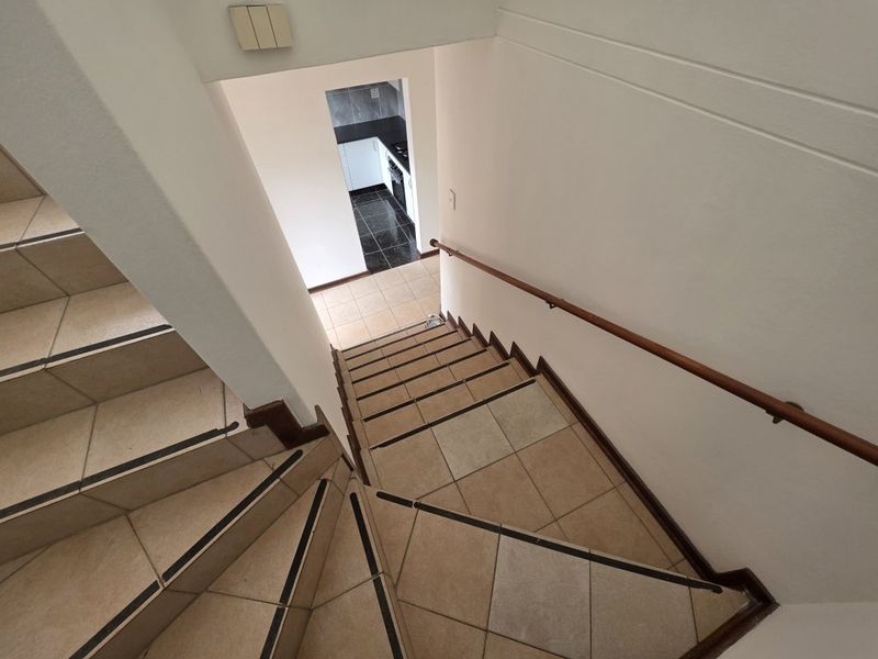 A view looking down a tiled staircase with a wooden handrail, leading towards a kitchen area with dark floor tiles.