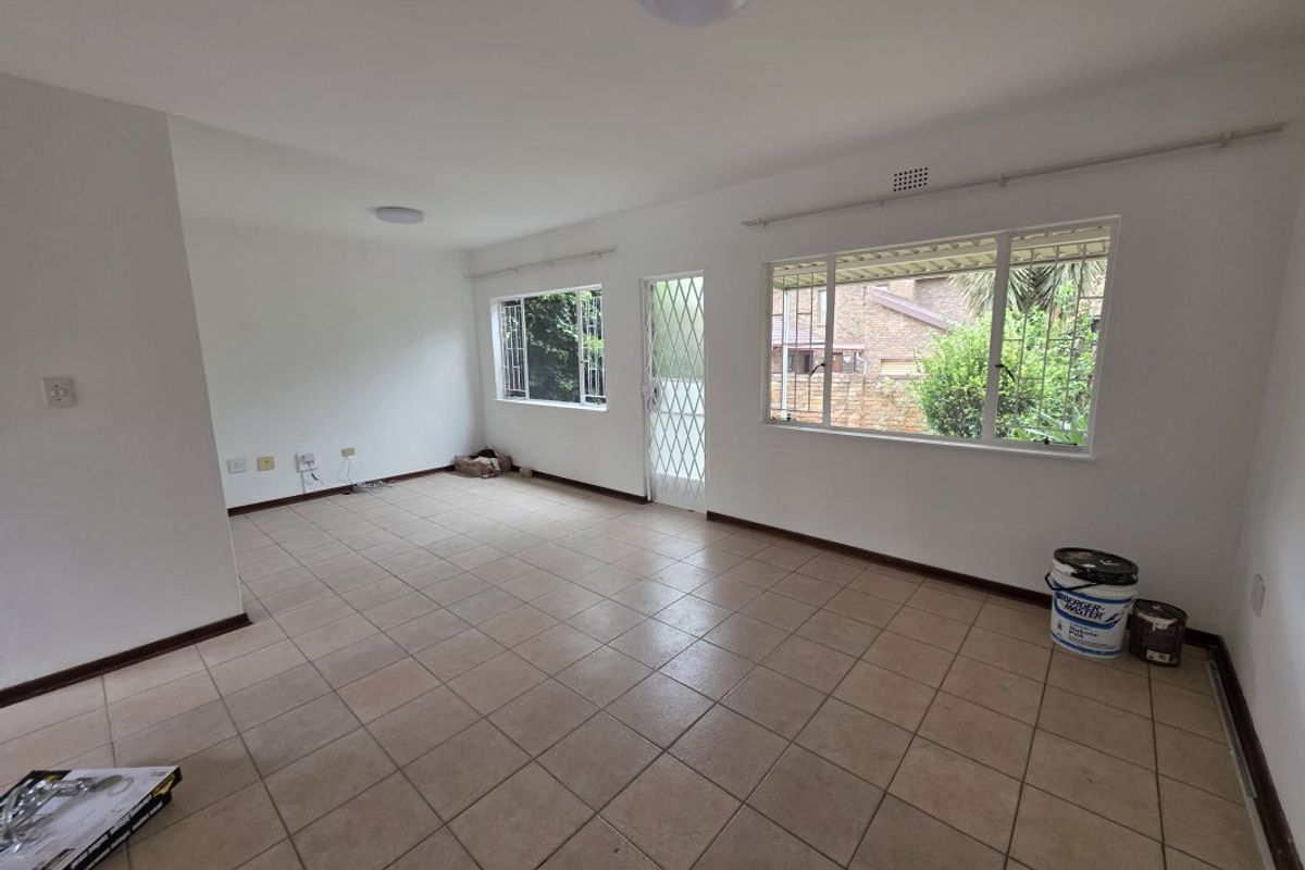 A spacious, empty living area featuring neutral tiled flooring, large windows with security bars, and a secure entrance door.
