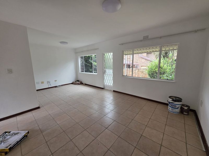 A spacious, empty living area featuring neutral tiled flooring, large windows with security bars, and a secure entrance door.