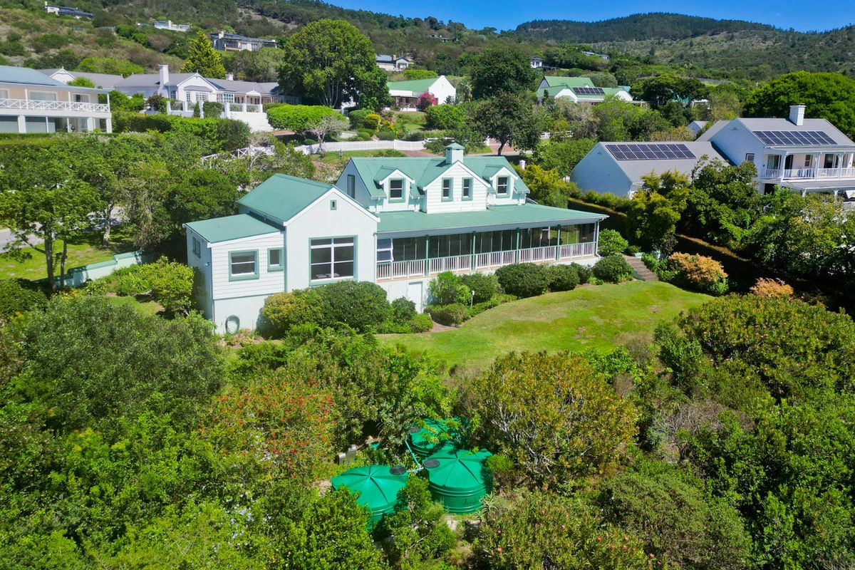Aerial view of a residential property featuring a green metal roof, wrap-around veranda, and lush garden with water storage tanks.
