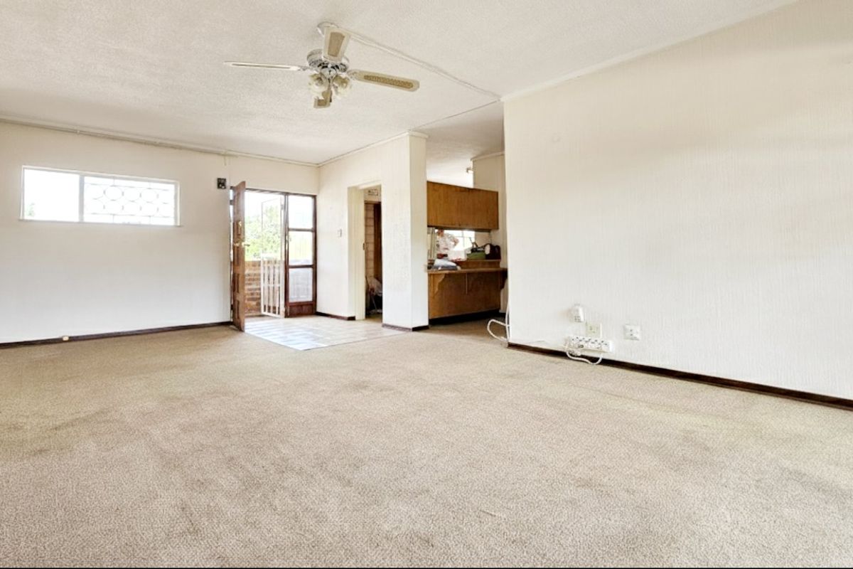 Spacious, empty living area featuring carpeted flooring, a ceiling fan, and an open-plan layout leading to a kitchen area with dated cabinetry.