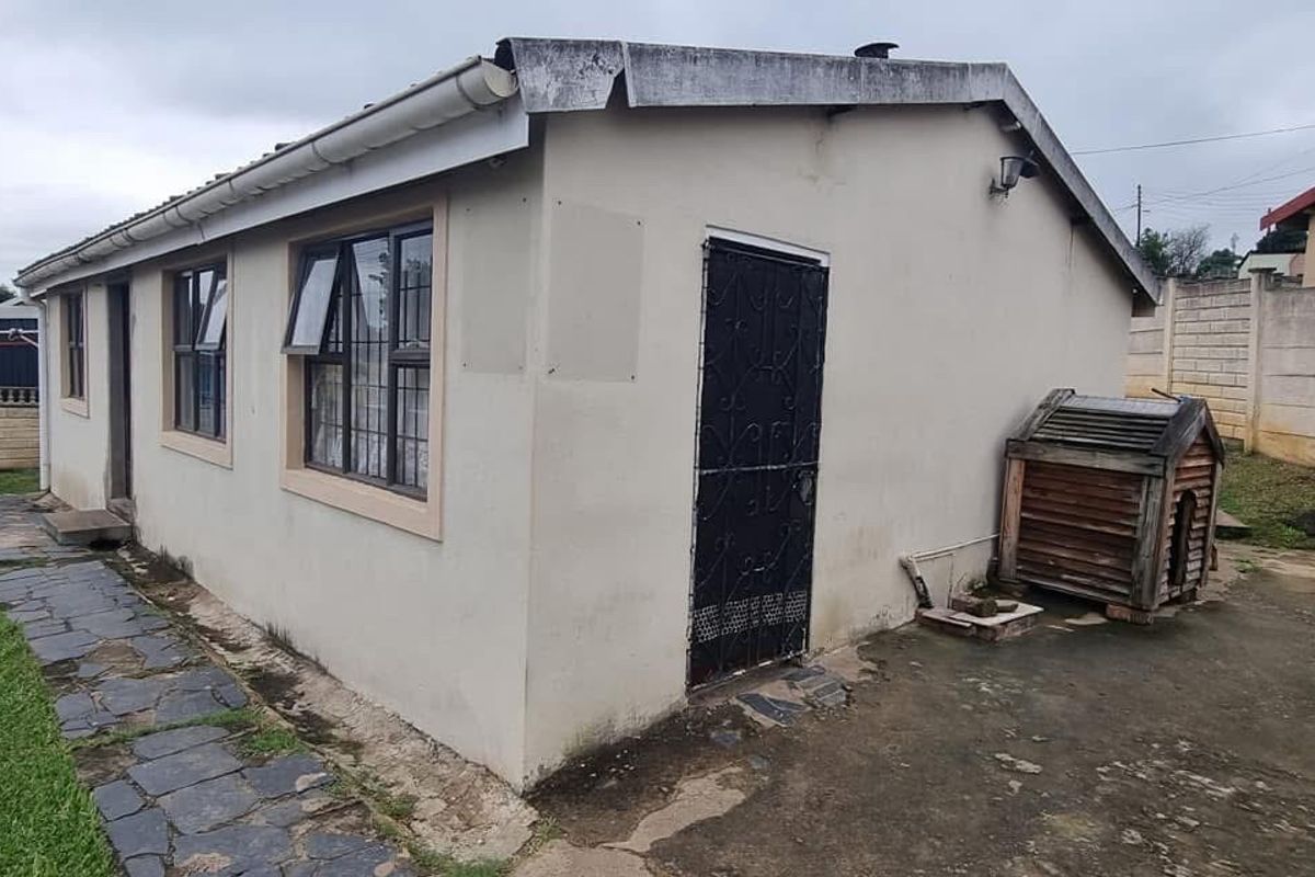 Rear exterior view of the property featuring a simple facade, security gate, aluminium windows, and a paved area enclosed by a precast boundary wall.