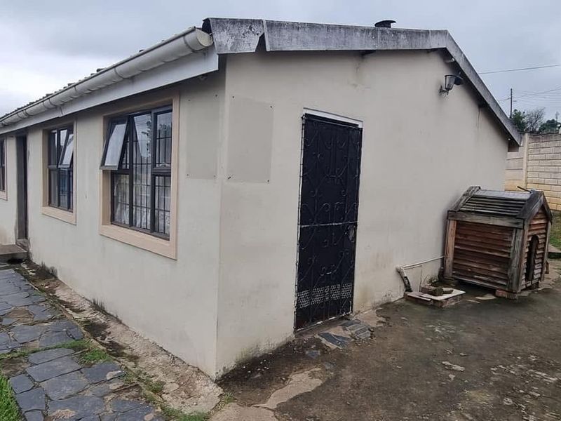 Rear exterior view of the property featuring a simple facade, security gate, aluminium windows, and a paved area enclosed by a precast boundary wall.