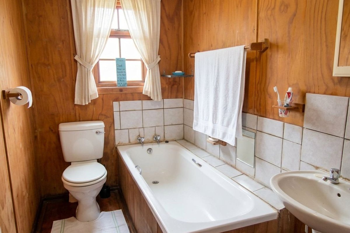 A compact bathroom featuring plywood wall paneling, a standard bathtub with tiled splashback, a pedestal sink, and a toilet.