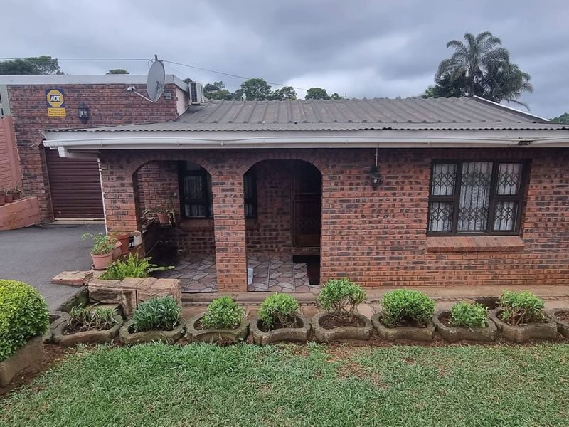 A single-story face-brick house featuring a covered front porch, metal roof, and a paved driveway with an adjacent garden area.