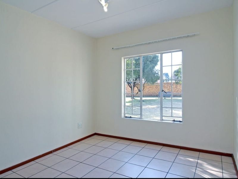 A bright, empty bedroom featuring neutral tiled flooring, clean walls, and a window with security bars overlooking the exterior grounds.