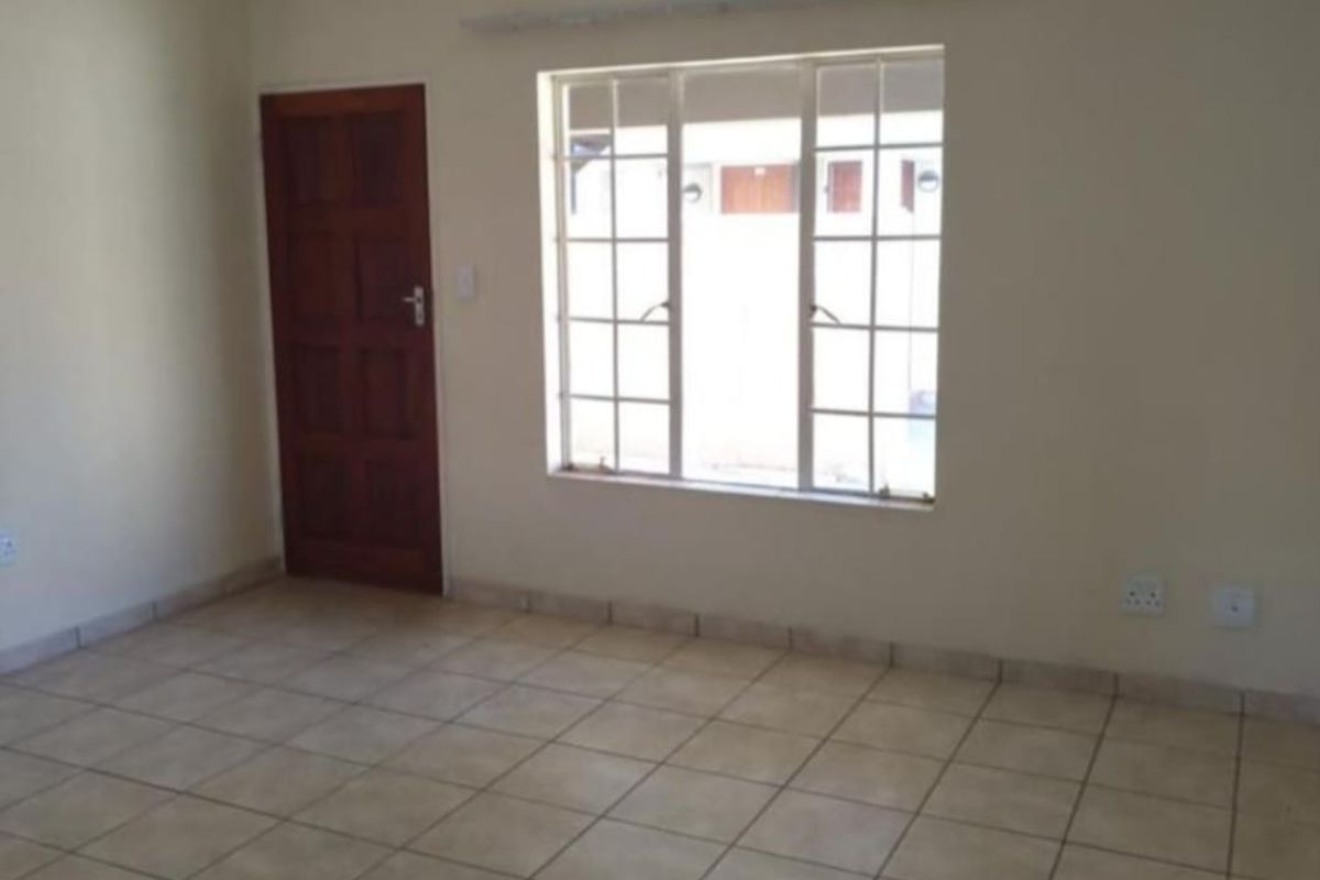 An empty living room featuring neutral tiled flooring, a wooden door, and a large grid-pattern window providing natural light.