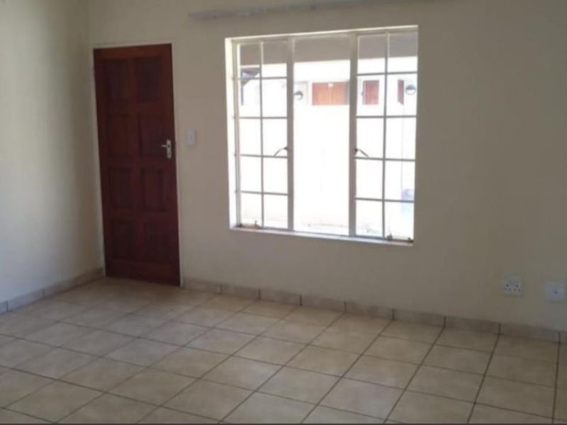 An empty living room featuring neutral tiled flooring, a wooden door, and a large grid-pattern window providing natural light.