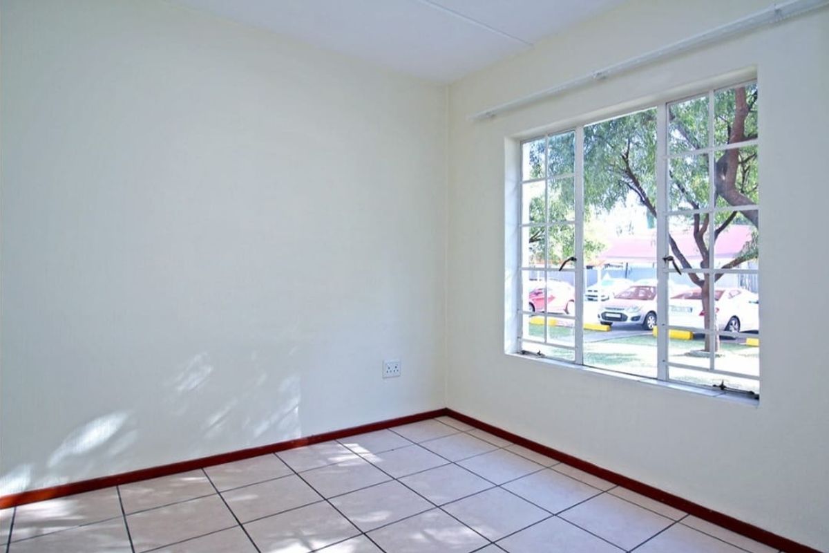 A bright, empty bedroom featuring neutral walls, tiled flooring, and a large window with security bars overlooking the exterior.