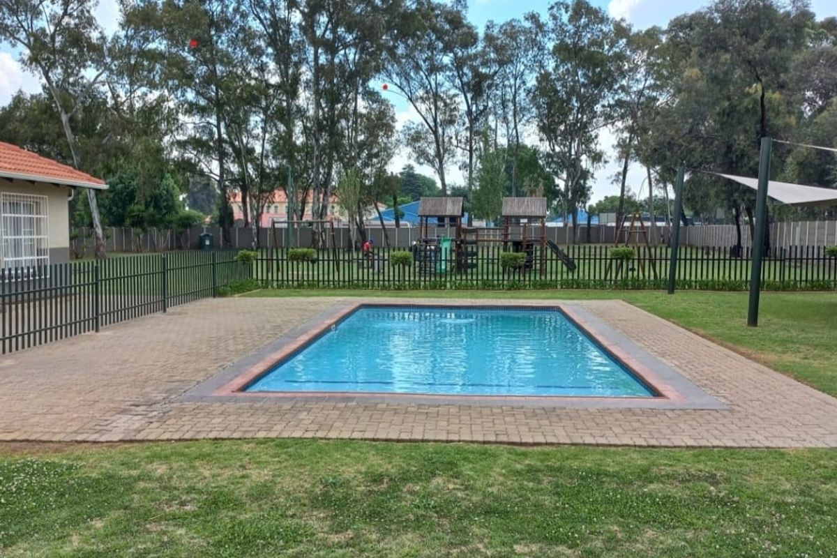 A communal outdoor area featuring a swimming pool, paved surround, and a fenced playground set within a landscaped garden.