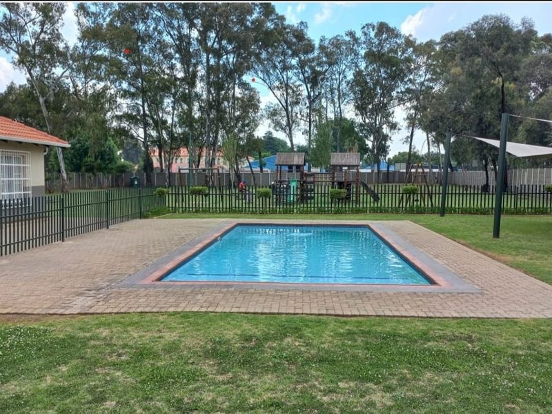 A communal outdoor area featuring a swimming pool, paved surround, and a fenced playground set within a landscaped garden.
