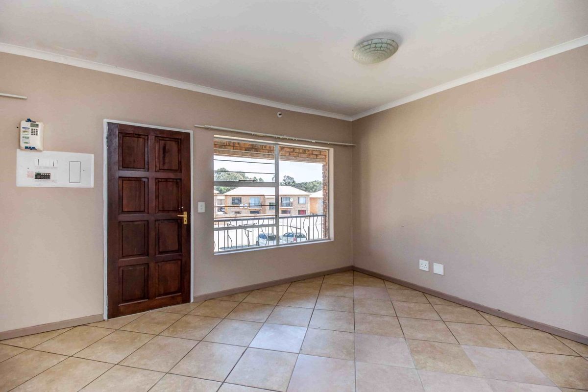 An empty living room featuring tiled flooring, a timber entrance door, and a large window providing natural light and views of the complex.