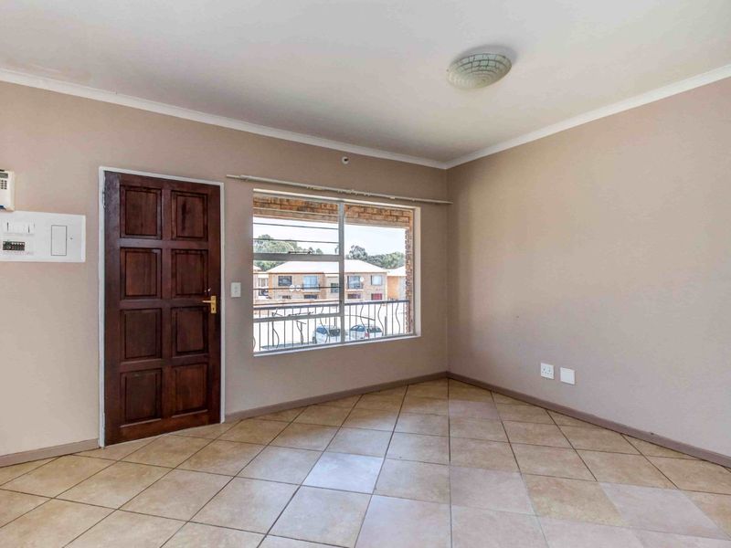 An empty living room featuring tiled flooring, a timber entrance door, and a large window providing natural light and views of the complex.