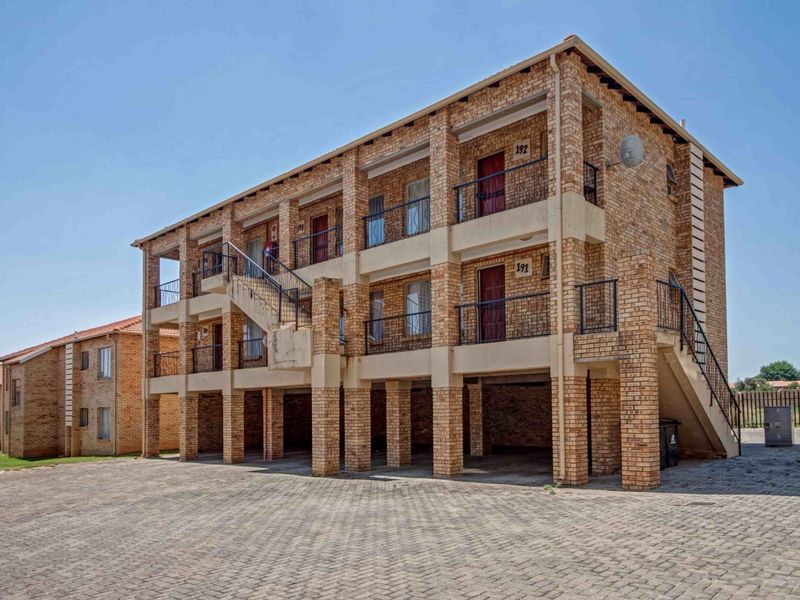 A multi-story apartment building featuring face brick construction, external staircases, private balconies, and a paved ground-level covered parking area.