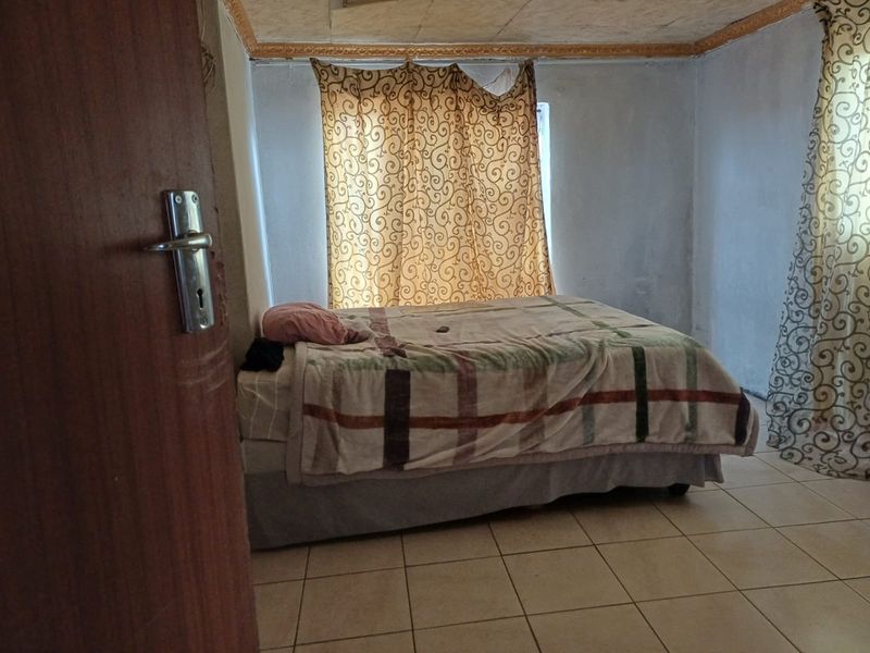 A bedroom featuring tiled flooring and a window with curtains, showing signs of wear on the walls and ceiling.