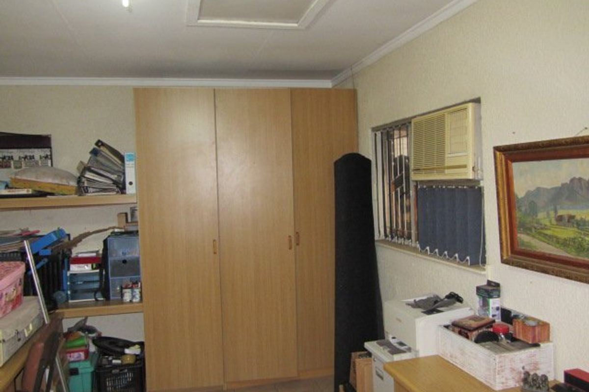 A bedroom featuring built-in wooden cupboards, tiled flooring, and a window-mounted air conditioning unit with security bars.