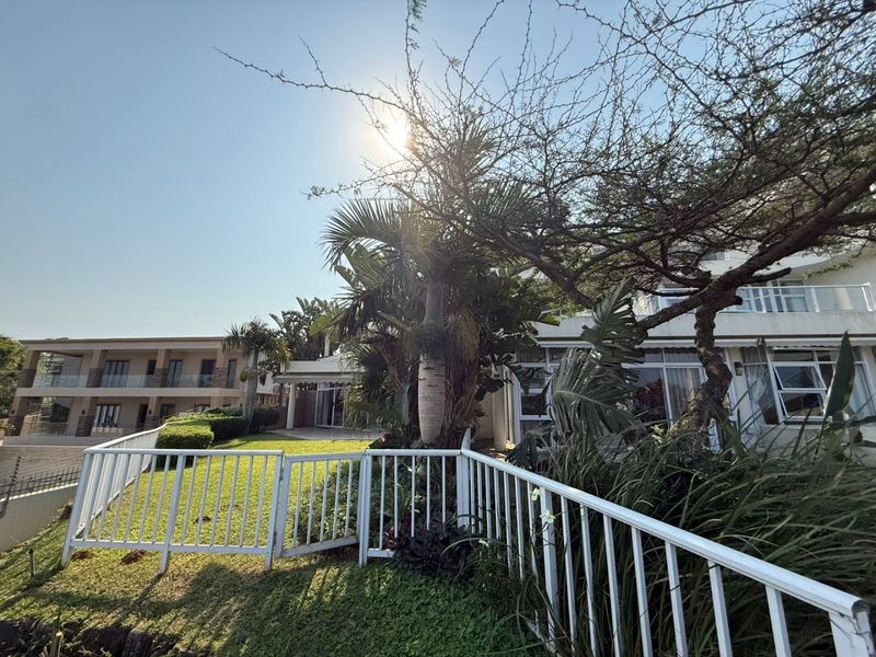 A view of the rear exterior of the apartment complex, featuring a well-maintained garden, white metal fencing, and a paved patio area.