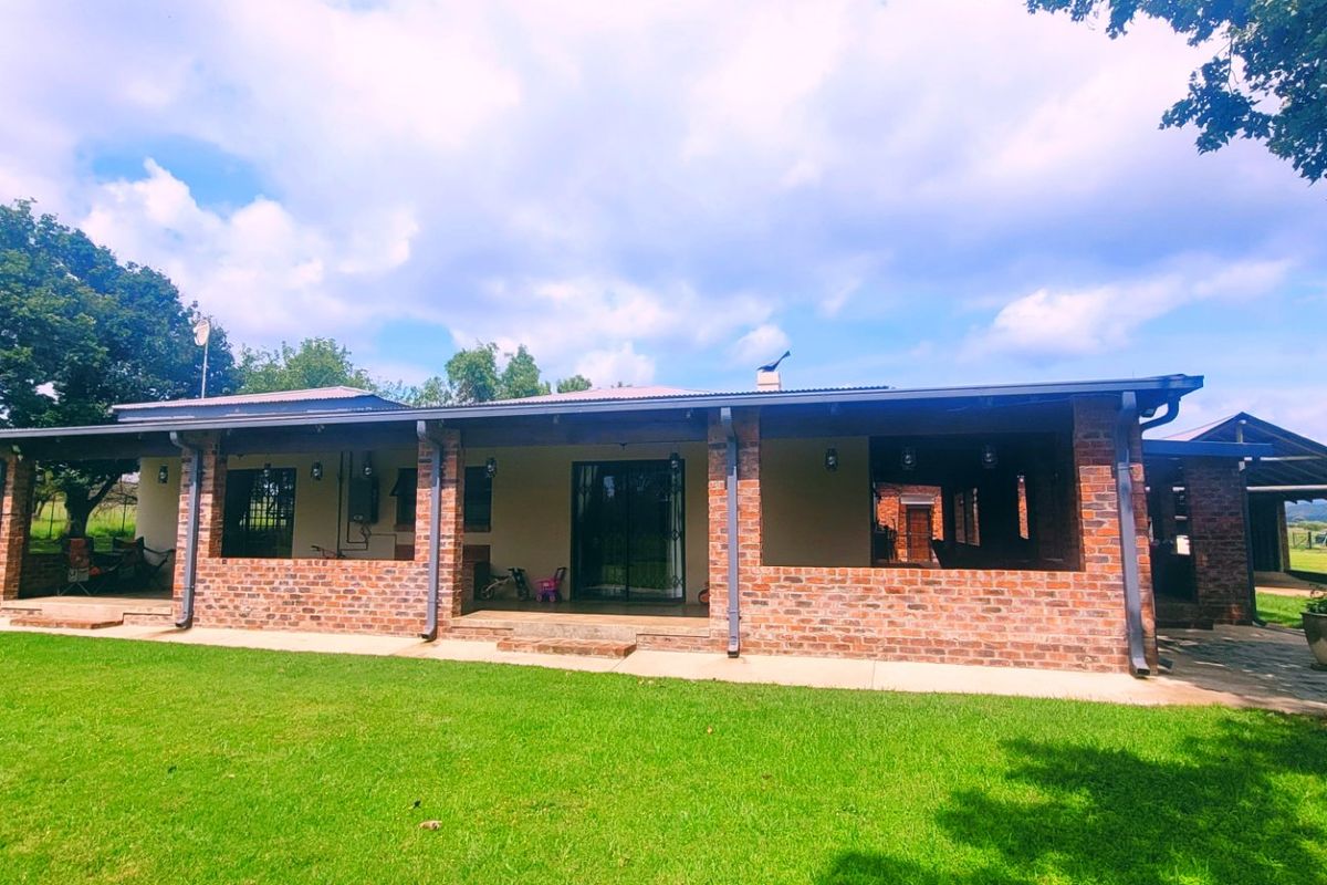 A well-maintained farmhouse exterior featuring an expansive covered brick patio, metal roofing, and large sliding glass doors overlooking a spacious green lawn.