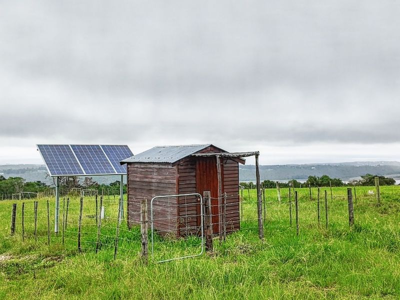 A small timber utility shed situated in a rural field, featuring an adjacent solar panel array and perimeter wire fencing.