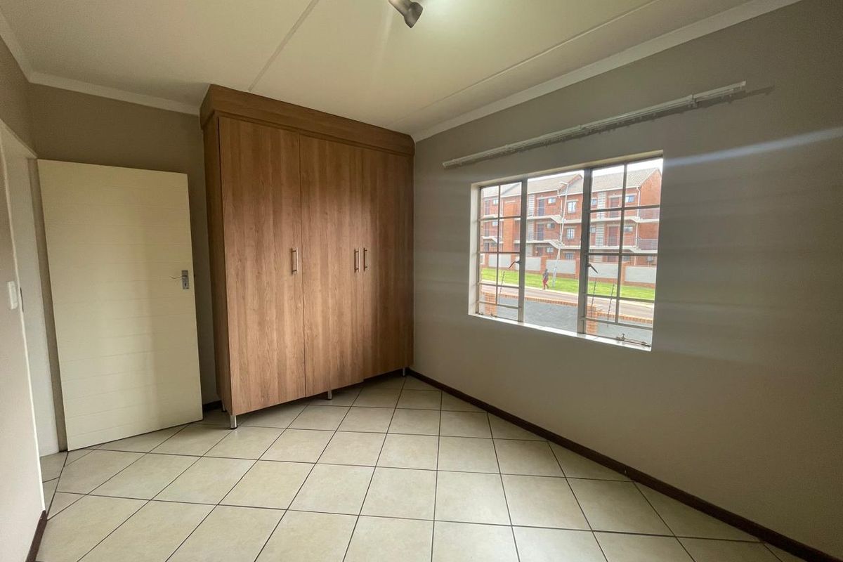 A bright, empty bedroom featuring neutral tiled flooring, a built-in wooden wardrobe, and a window with security bars.