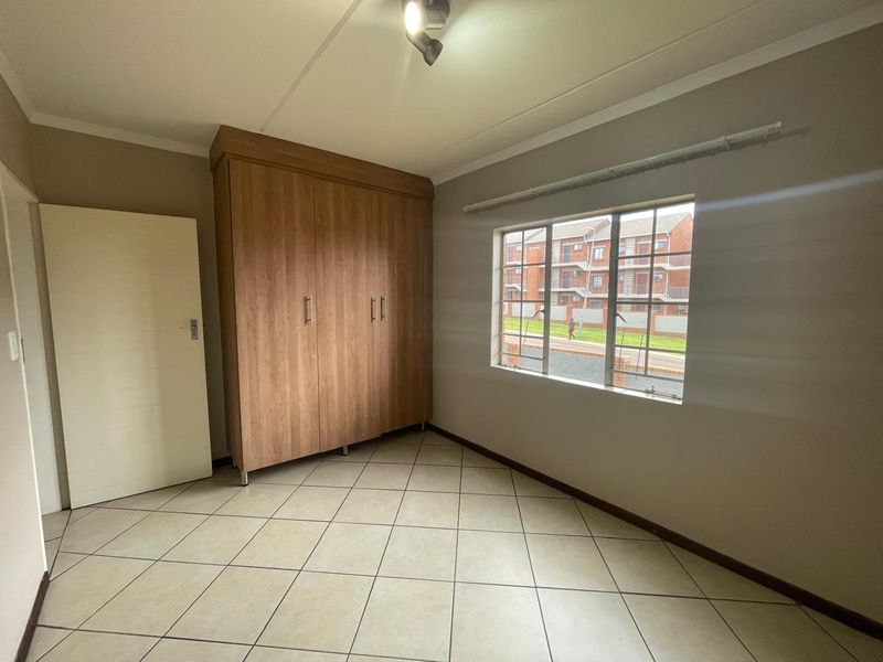 A bright, empty bedroom featuring neutral tiled flooring, a built-in wooden wardrobe, and a window with security bars.