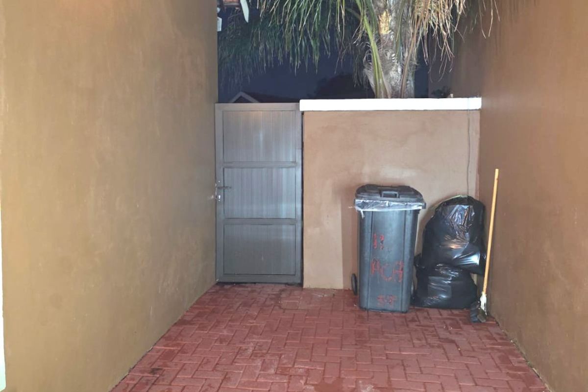 A narrow exterior side passage featuring red brick paving, a secure metal gate, and an outdoor wall-mounted light fixture.