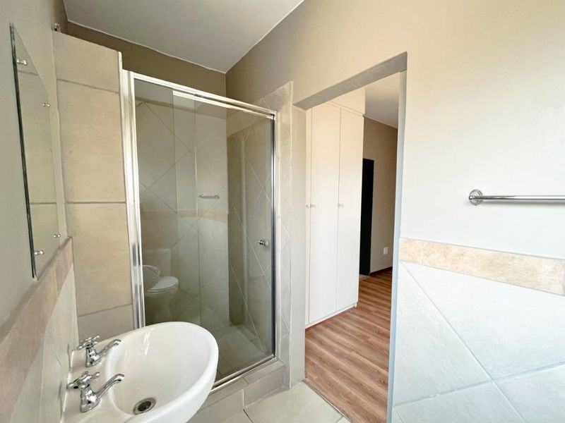 A clean, functional bathroom featuring a glass-enclosed shower, pedestal basin, and neutral wall tiling, with adjacent built-in storage cupboards.