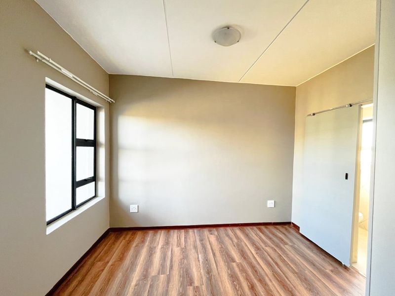 A bright, empty bedroom featuring modern laminate flooring, neutral wall tones, and a sliding door leading to an adjacent space.
